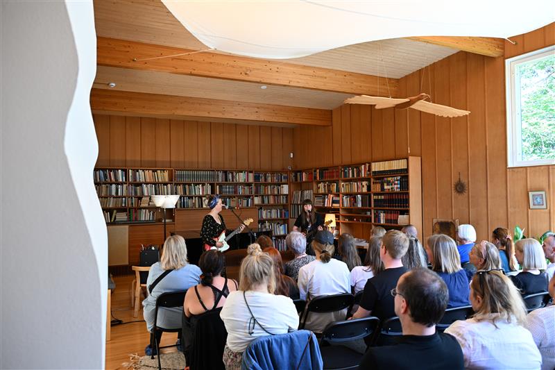 Ihmiset istuvat huoneessa, jossa on puupaneeliseinät ja kirjahyllyjä. Kaksi muusikkoa esiintyy ihmisille.